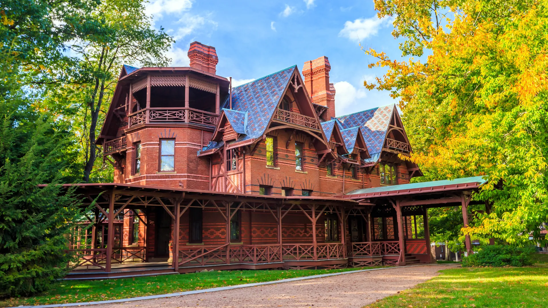 A large, ornate red-brick mansion stands surrounded by lush green trees and blue skies