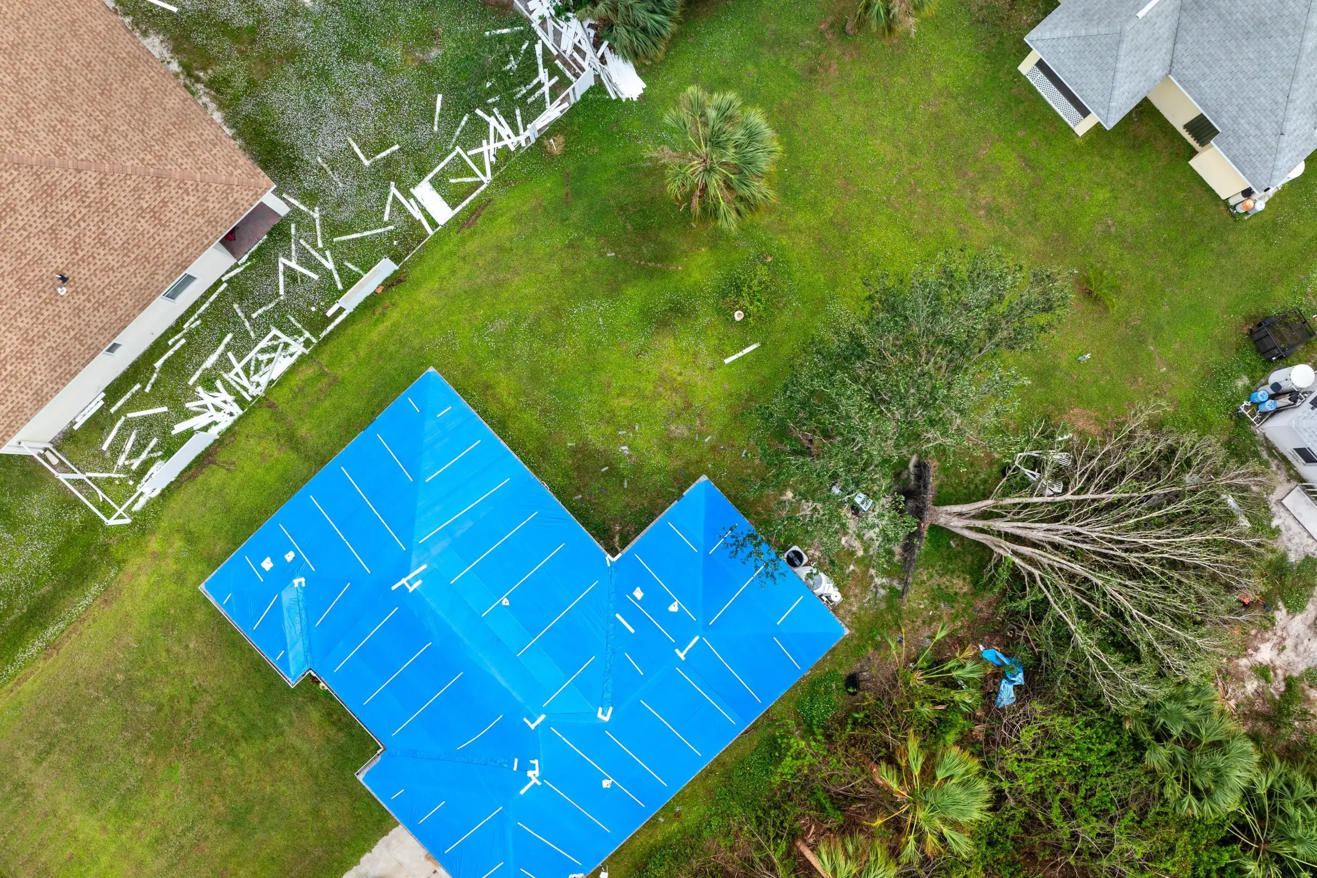 A blue pool cover sits on a yard with scattered debris and a fallen tree